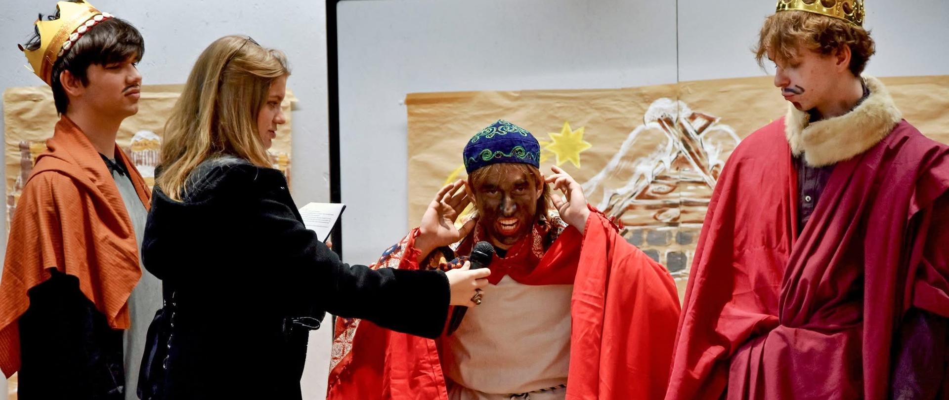 Trzy młode osoby w przebraniach trzech mędrców (królów), na tle ręcznie malowanych dekoracji. Pomiędzy nimi młoda osoba w eleganckim płaszczu, trzyma mikrofon przy głowie jednej z przebranych postaci