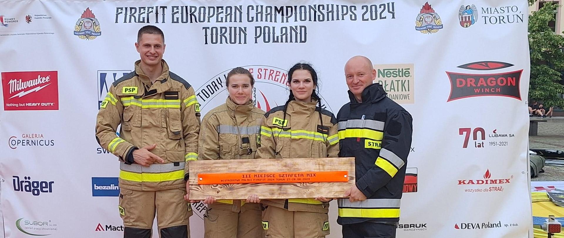 Pamiątkowe zdjęcie na ściance. Reprezentacja SA PSP w Krakowie ze zdobytym trofeum zawodów TFA – cztery osoby w umundurowaniu specjalnym. 