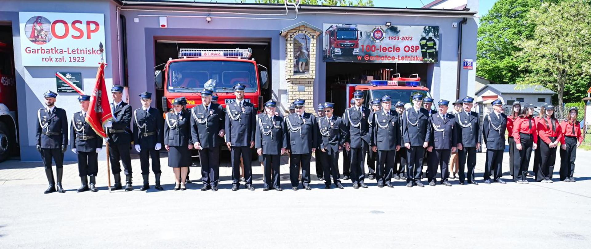 strażacy ochotnicy stoją na placu przed remizą osp ubrani w mundury, w tle samochody strażackie w otwartych garażach