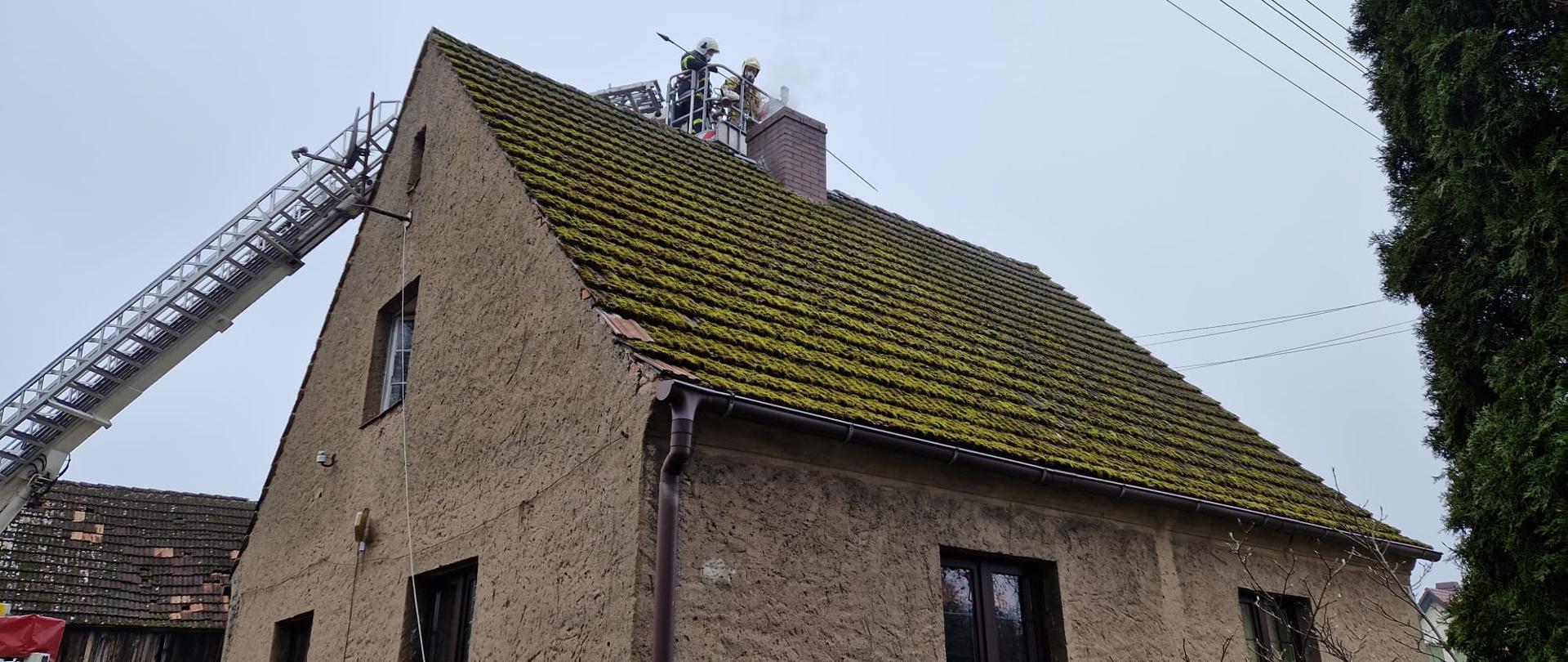 Fotografia przedstawia budynek mieszkalny jednorodzinny z dachem prostym dwuspadowym, przy budynku rozstawiony podnośnik, na którym strażacy prowadzą działania przy kominie
