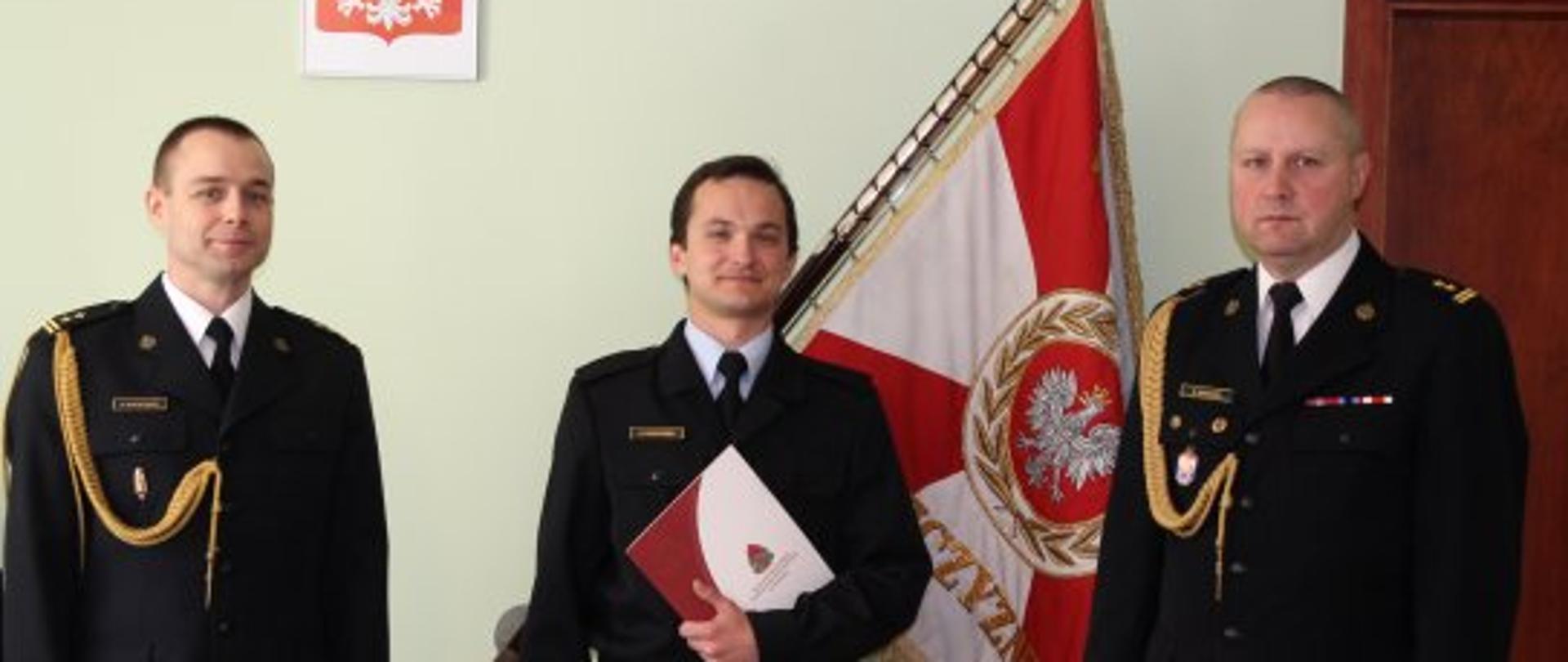 na jasnej ścianie stoi trzech strażaków ubranych w ciemne mundury w tle widać sztandar oraz godło