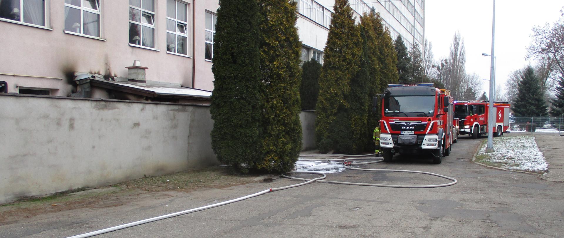 Na zdjęciu widać dwa wozy bojowe straży pożarnej. Posiadają sygnalizację ostrzegawczą, mają oznakowanie. Stoją przed budynkiem. Obok aut krzewy ozdobne. Budynek szary. Niebo zachmurzone. Auta stoją na szarym asfalcie. Widać białe linie wężowe.