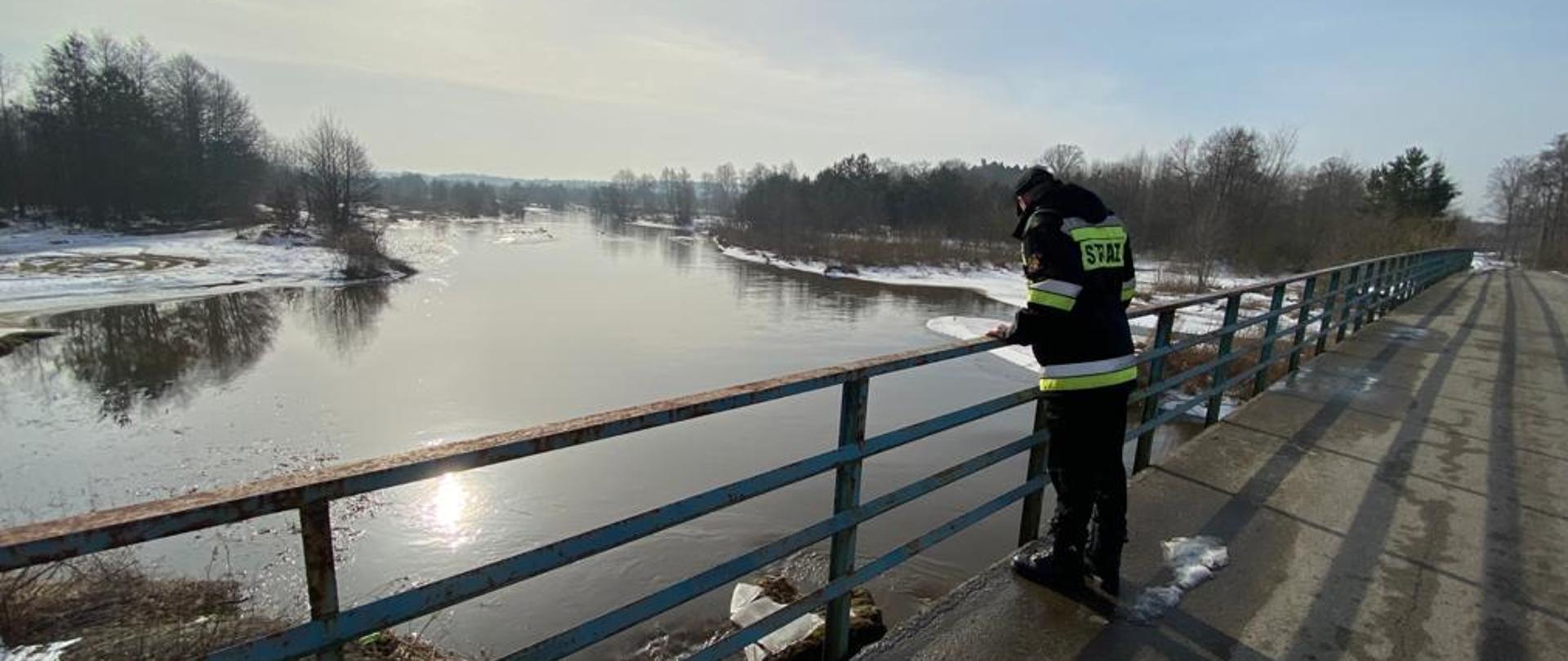 Strażak stojący na moście. Strażak obserwuje przepływającą poniżej rzekę. Rzeka ma wysoki poziom woda i ma szerokie koryto. 