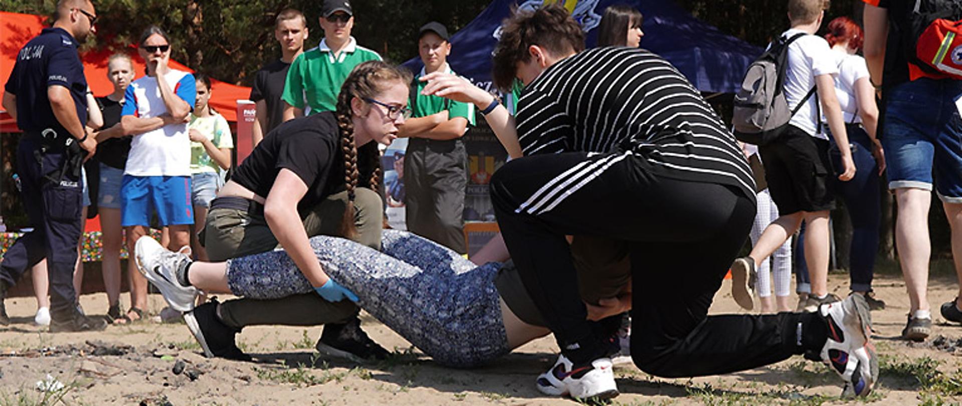 Zdjęcie przedstawia młoda dziewczynę i młodego chłopaka udzielających pomoc osobie. Dziewczyna ma na dłoniach rękawiczki lateksowe, trzyma osobę poszkodowaną za nogi, chłopak trzyma poszkodowanego za tułów od strony głowy. W tle osoby przyglądające się akcji ratunkowej.