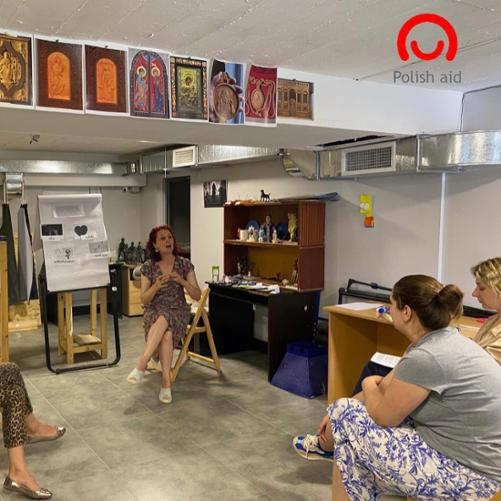 W sali kilka kobiet siedzi na ustawionych w półkolu krzesłach i słuchają prowadzącej szkolenie