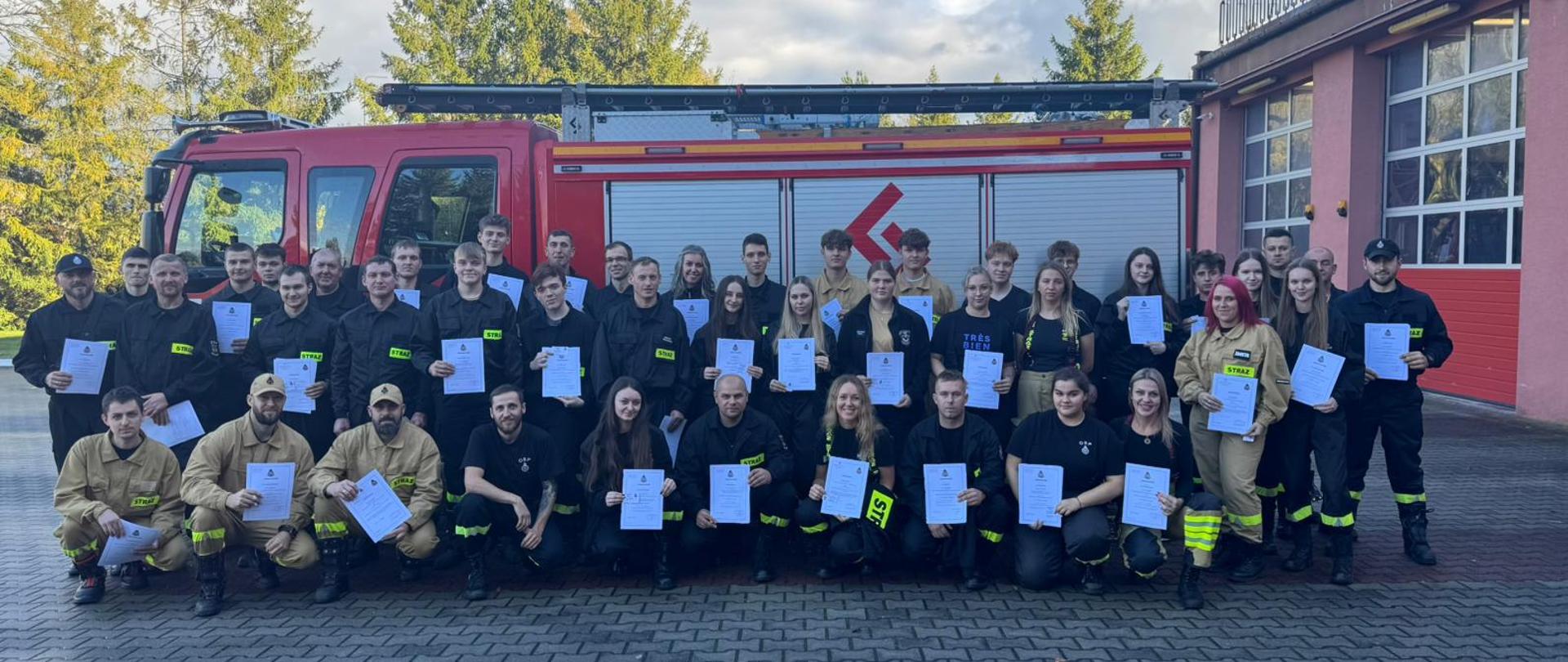 na zdjęciu widoczni strażacy OSP z dyplomami ukończenia kursu na tle samochodu ciężarowego straży w oddali drzewa i niebo po prawo budynek straży pożarnej