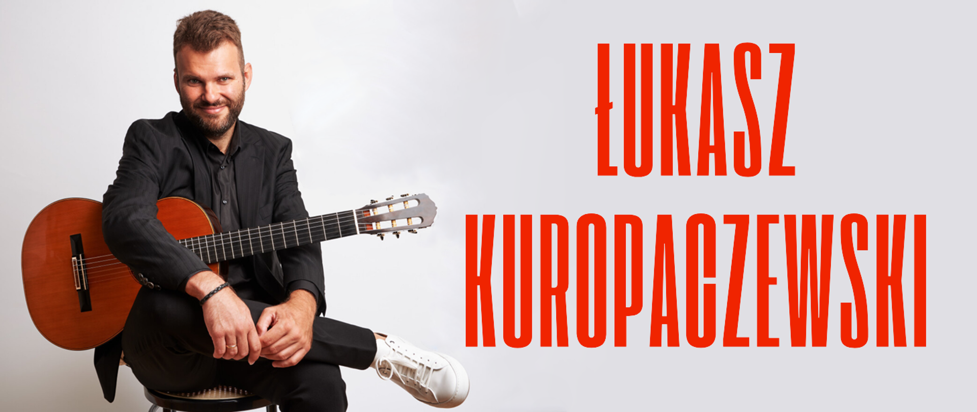 Baner przedstawia zdjęcie siedzącego gitarzysty z założoną nogą. Ubrany jest w czarny garnitur, a na nogach trzyma gitarę. Zdjęcie na szarym tle. Obok niego duży czerwony napis „Łukasz Kuropaczewski”.