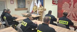 strażacy ubrani czarne ubrania koszarowe przed egzaminem teoretycznym w Sali szkoleniowej, w tle funkcjonariusze z komisje egzaminacyjnej ubrani w piaskowe mundury, ponadto godło i flaga Polski