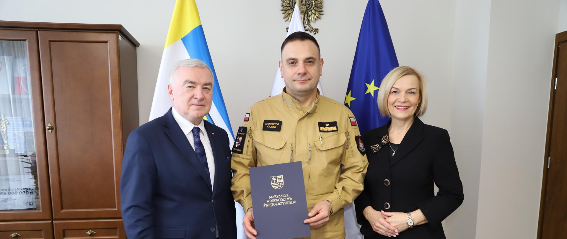 Na zdjęciu widzimy od lewej marszałka województwa świętokrzyskiego w garniturze, w środku strażaka generała w mundurze piaskowym trzymającego w ręku teczkę z napisem "Marszałek Województwa Świętokrzyskiego" oraz z prawej kobietę w garsonce czarnej - wicemarszałek województwa. W tle flagi.