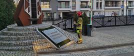 Strażak Państwowej Straży Pożarnej trzyma znicz przed pomnikiem "W hołdzie pomorskim strażakom". 