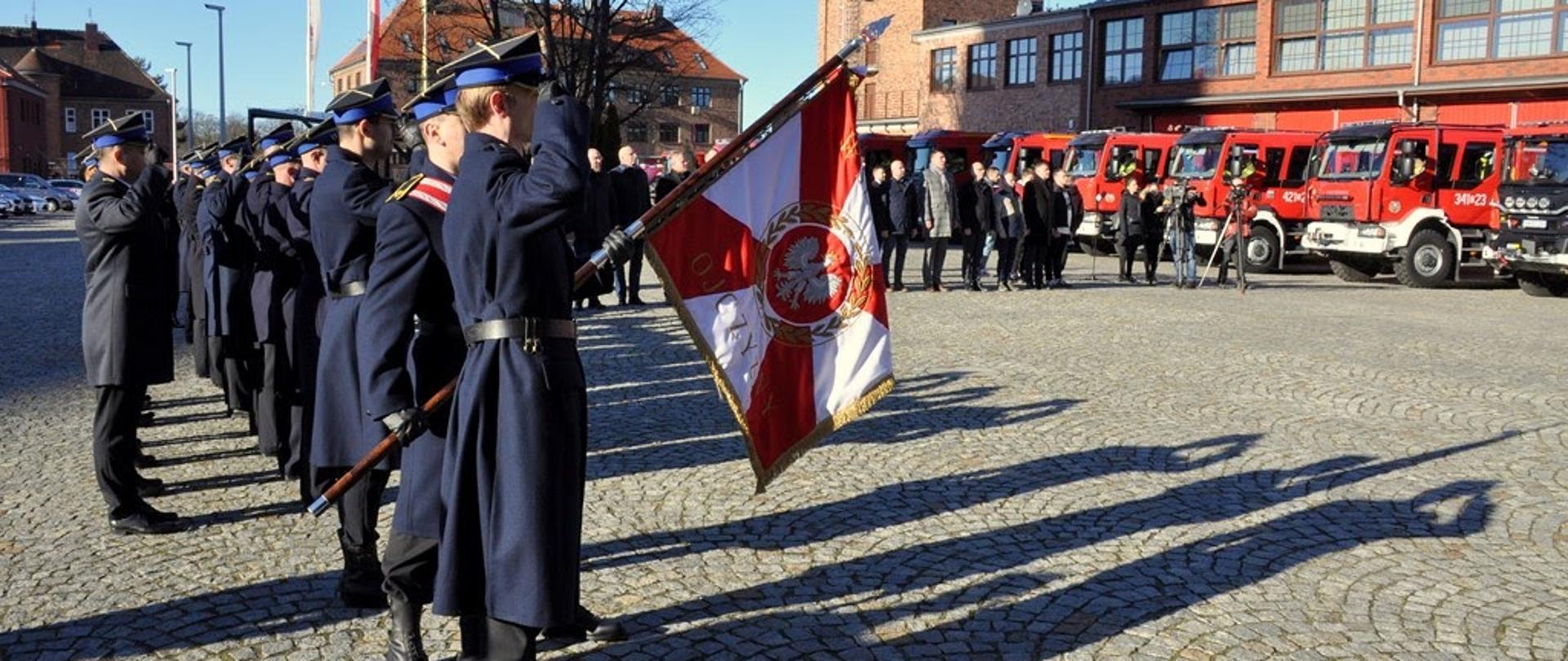 Widok na wewnętrzny plac Komendy Wojewódzkiej we Wrocławiu. Po prawej stronie przed bramami garażwymi w rzędzie stoją pojazdy ratowniczo-gaśnicze. Po prawej stronie stoją funkcjonariusze w umundurowaniu galowym oraz poczet sztandarowy.