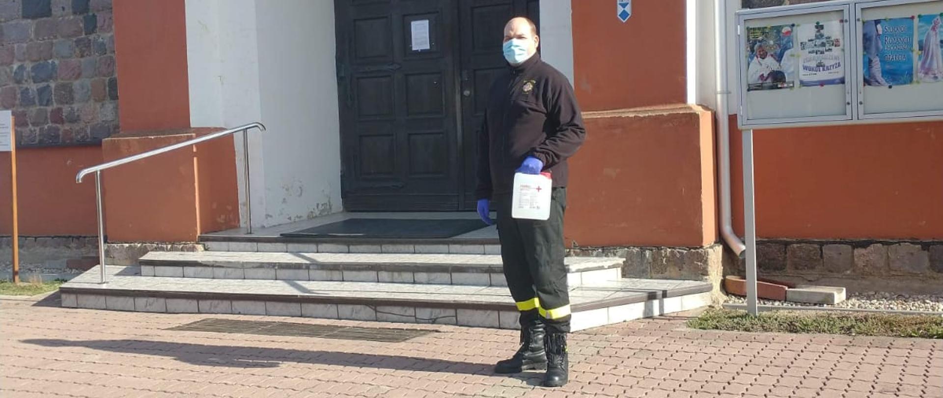 Na zdjęciu widoczny strażak przed budynkiem kościoła podczas dostarczenia płynu dezynfekcyjnego. 