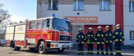 Widok z przodu. Pięciu strażaków OSP Zakrzów stoi w ubraniach specjalnych przed samochodem pożarniczym przed garażem OSP. 