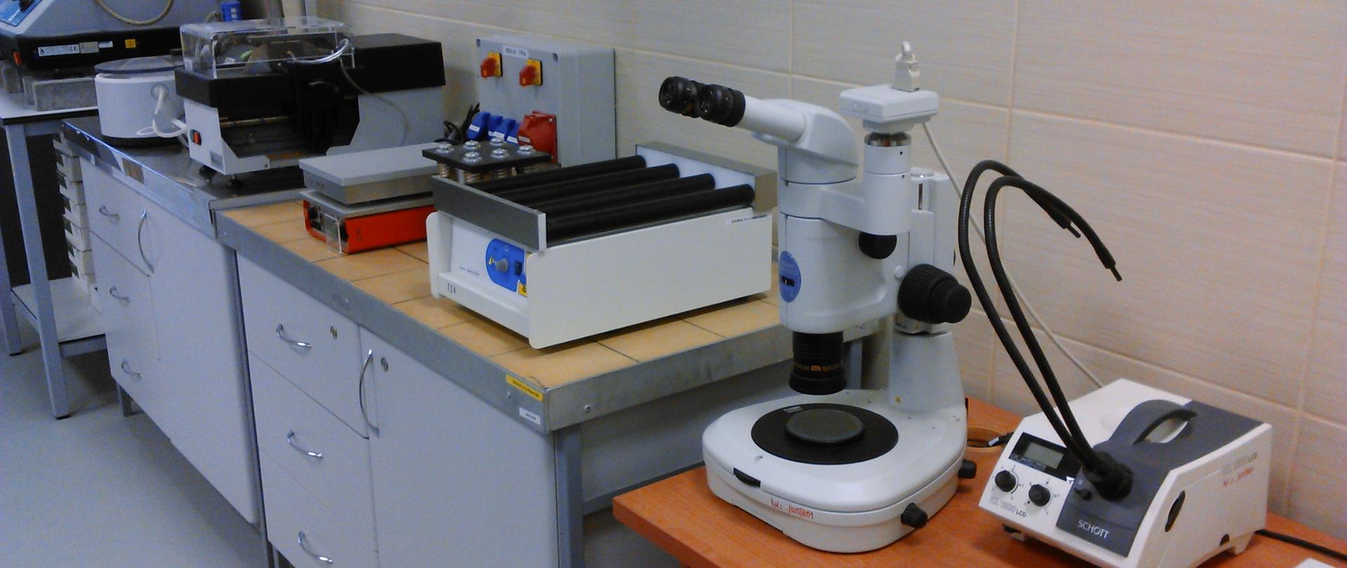Stanowisko laboratoryjne do badań petrograficznych kruszyw. Na blacie znajdują się różne urządzenia: stereomikroskop z oświetleniem, mieszadło rolkowe, waga laboratoryjna oraz inne przyrządy analityczne. Sprzęt ustawiony jest na szafkach i stole w jasnym pomieszczeniu z płytkami na ścianie.