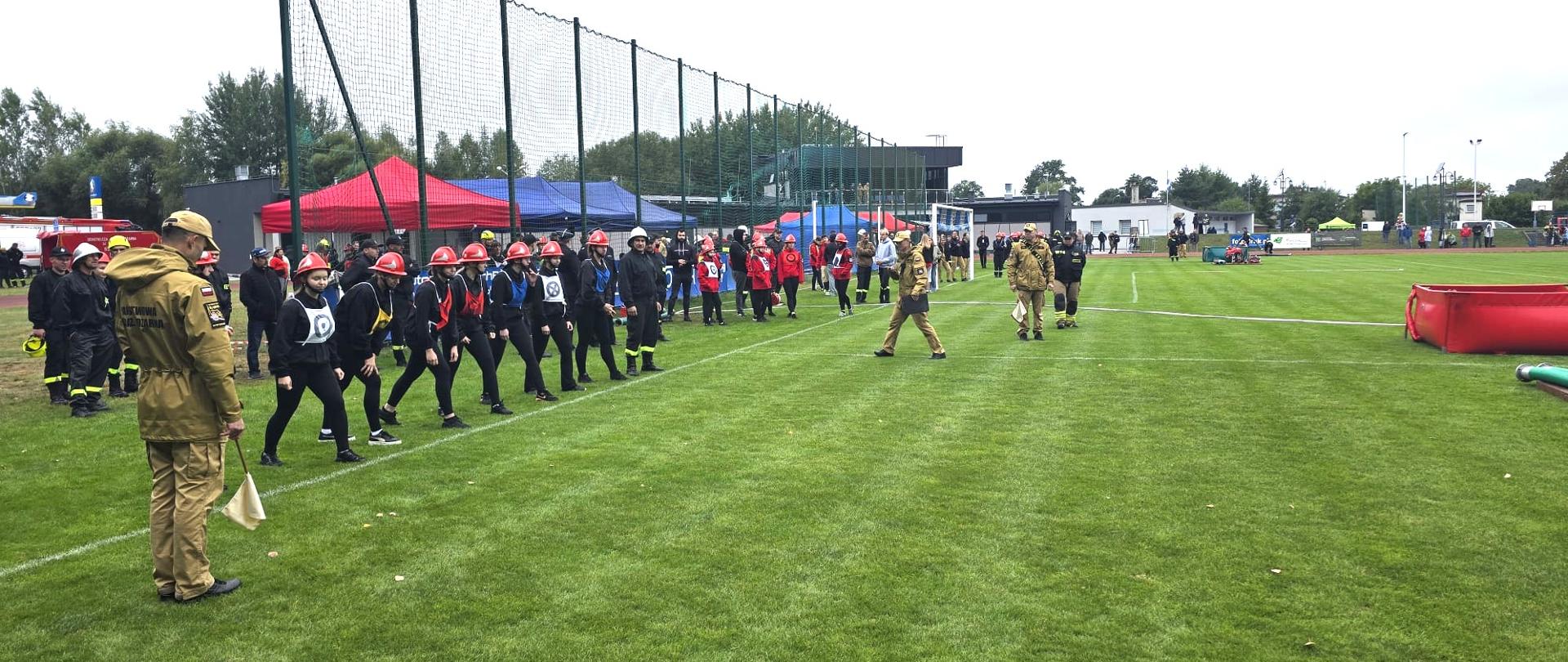 Na zdjęciu widać grupę młodych ludzi w czarnych strojach i czerwonych kaskach, stojących w rzędzie na trawiastym boisku. Obok nich znajdują się dorośli sędziowie w mundurach. W tle widać trybuny, siatki zabezpieczające oraz czerwone namioty. Wygląda na to, że są to zawody.