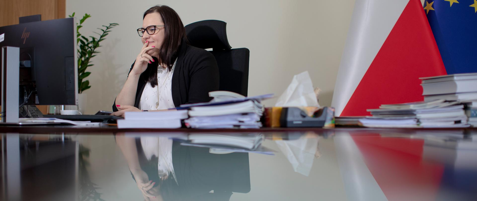Wiceminister Małgorzata Jarosińska-Jedynak siedzi przy biurku w gabinecie przed monitorem komputera. Za nią flagi PL i UE.