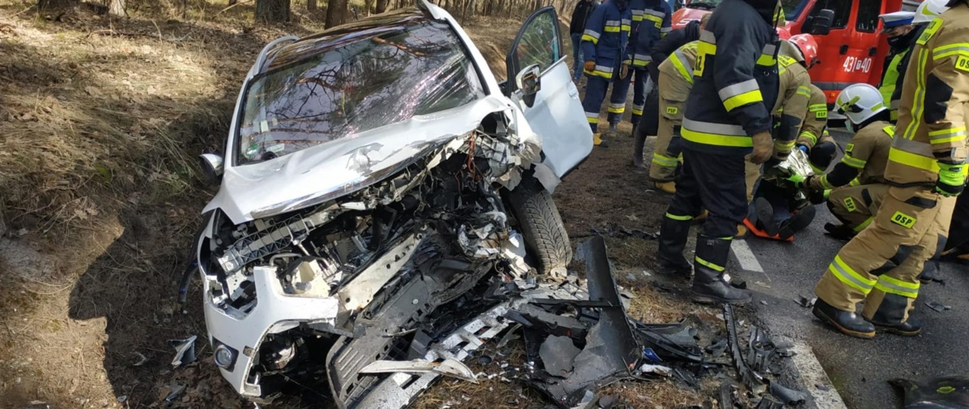 Po lewej - rozbity przód samochodu osobowego stojącego w rowie, po prawej strażacy OSP i PSP prowadzą działania.