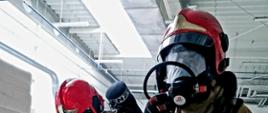 Strażacy ubrani w piaskowe ubranie specjalne i czerwony hełm w hali produkcyjnej ewakuują manekina.