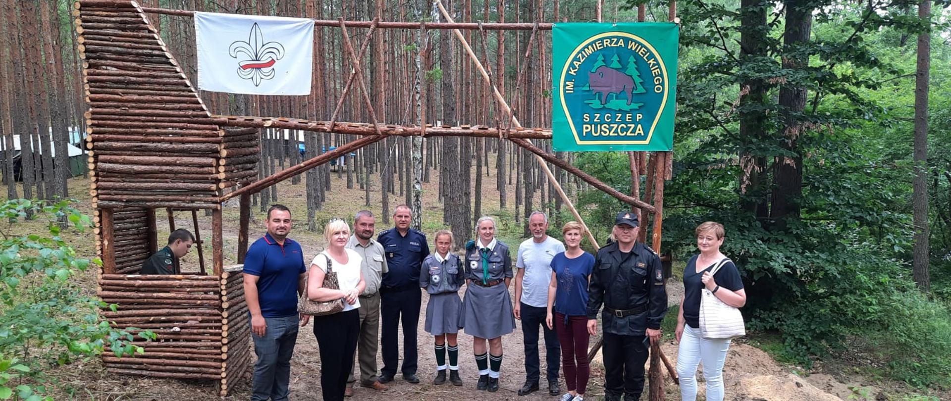 Fotografa przedstawia ujęcie bramy do obozu harcerskiego. Brama wykonana jest z sosnowych żerdzi. Na bramie zawieszone są proporce Związku Harcerstwa Rzeczypospolitej oraz Szczepu „Puszcza”. Na tle bramy stoją osoby wizytujące obóz harcerski oraz pośrodku tej grupy dwie harcerki. W tle las sosnowy. Jest dzień, słonecznie.