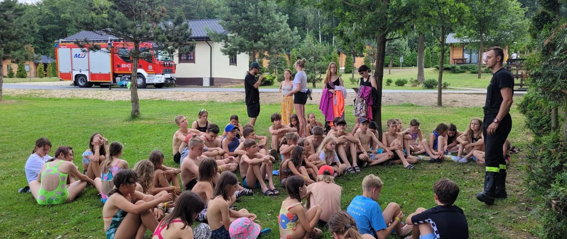 Zdjęcie przedstawia dużą grupę dzieci siedzących na trawie w półkolu, prawdopodobnie podczas letniego obozu lub spotkania. Większość dzieci ubrana jest w stroje kąpielowe, co sugeruje aktywności wodne lub upalną pogodę. W tle widocznych jest kilku dorosłych, w tym jeden mężczyzna w mundurze strażackim, oraz czerwony wóz strażacki zaparkowany pod drzewami. Całość rozgrywa się na terenie zielonym, otoczonym drzewami i widać fragmenty budynków w tle.