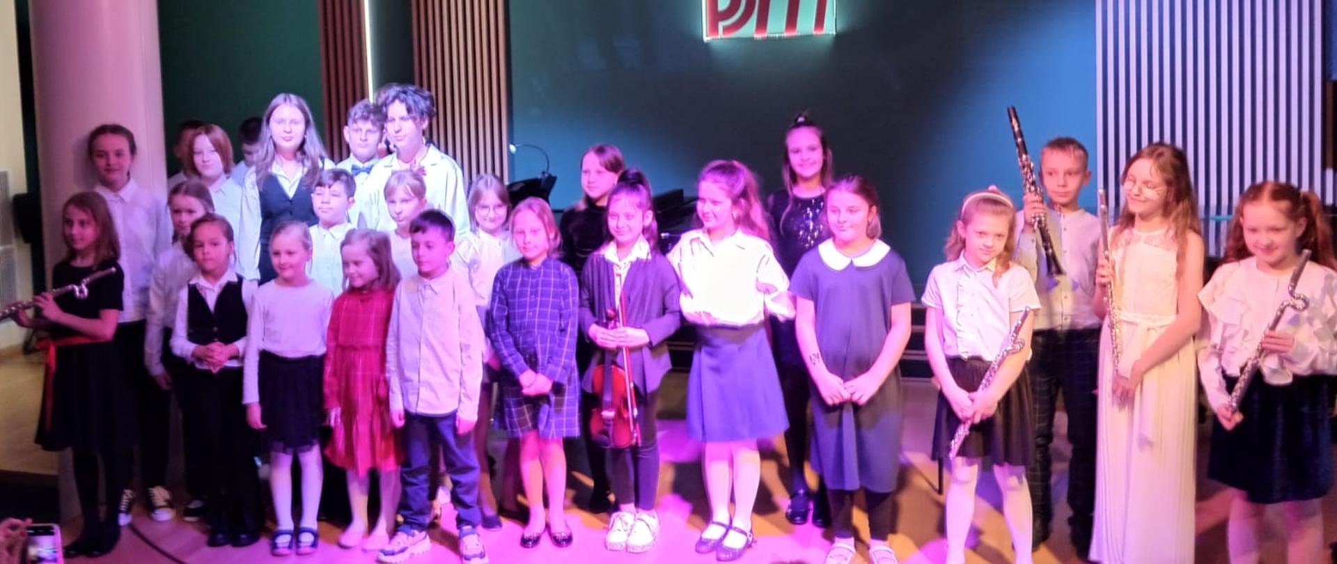 Planie na zdjęciu widoczni są uczniowie którzy stoją w galowych strojach koncertowych z instrumentami w rękach za nimi ciemnozielona ściana z nowo wyremontowanymi lamelami oraz fragmentem napisu logo szkoły PSM