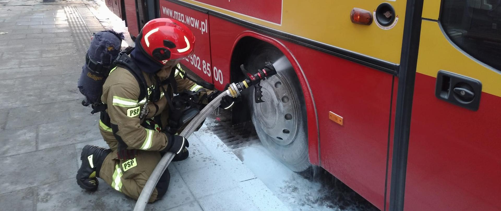 Na zdjęciu widać autobus koloru żółto- czerwonego w którym doszło do pożaru układu hamulcowego. Strażak w piaskowym ubraniu specjalnym wyposażony w aparat dróg oddechowych, który chłodzi powierzchnie elementów objęte pożarem