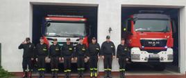 Zdjęcie przedstawia strażaków stojących przed garażem podczas oddawania hołdu zmarłemu strażakowi. Strażacy ubrani w ubrania koszarowe. Za nimi stoją samochody strażackie z włączonymi sygnałami świetlnymi. 