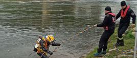 Na zdjęciu widać nurka ubranego w zestaw do nurkowania przewodowego z hełmem Kirby-Morgan podczas wychodzenia z wody na brzeg rzeki SAN w asyście dwóch asekurujących ratowników. W tle most kolejowy w Przemyślu