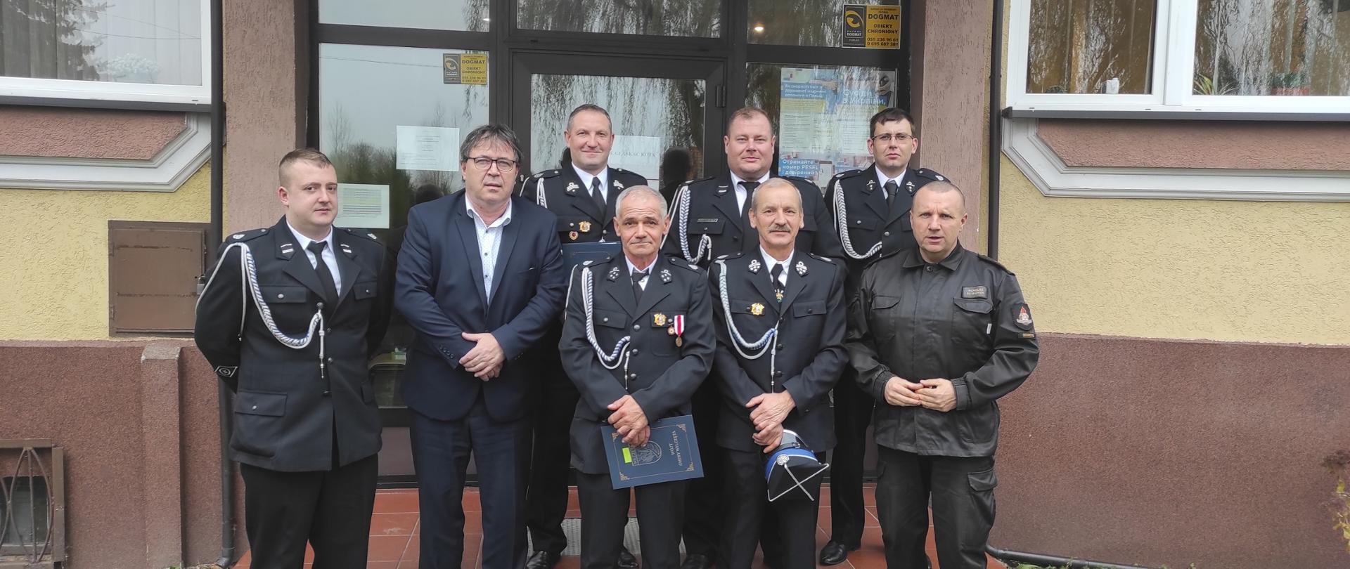 Przed wejściem do budynku urzędu do zdjęcia pozują, sześciu strażaków ochotników w mundurach wyjściowych, wójt oraz komendant powiatowy w czarnym mundurze.