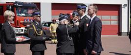 pięciu strażaków oraz jeden cywil stoją na placu komendy podczas ceremonii wręczania odznaczenia, przed nimi stoi strażak ubrany w mundur galowy strażaka ochotniczej straży pożarnej i dekoruje strażaka odznaką, za nim stoi zastępca łódzkiego komendanta wojewódzkiego oraz osoba cywilna trzymająca tacę z odznaczeniami, w tle budynek garażowy komendy oraz dwa pojazdy strażackie, pogoda słoneczna