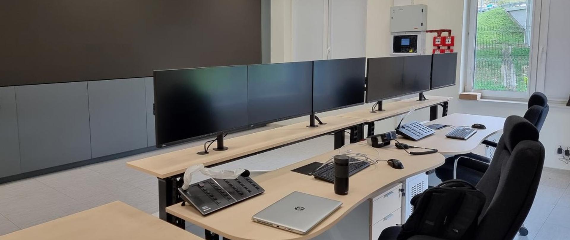 Pomieszczenie biurowe. Stanowisko w Centrum Zarządzania Tunelem. Jeden wielki ekran wiszący na ścianie i szereg mniejszych ustawionych na biurku.