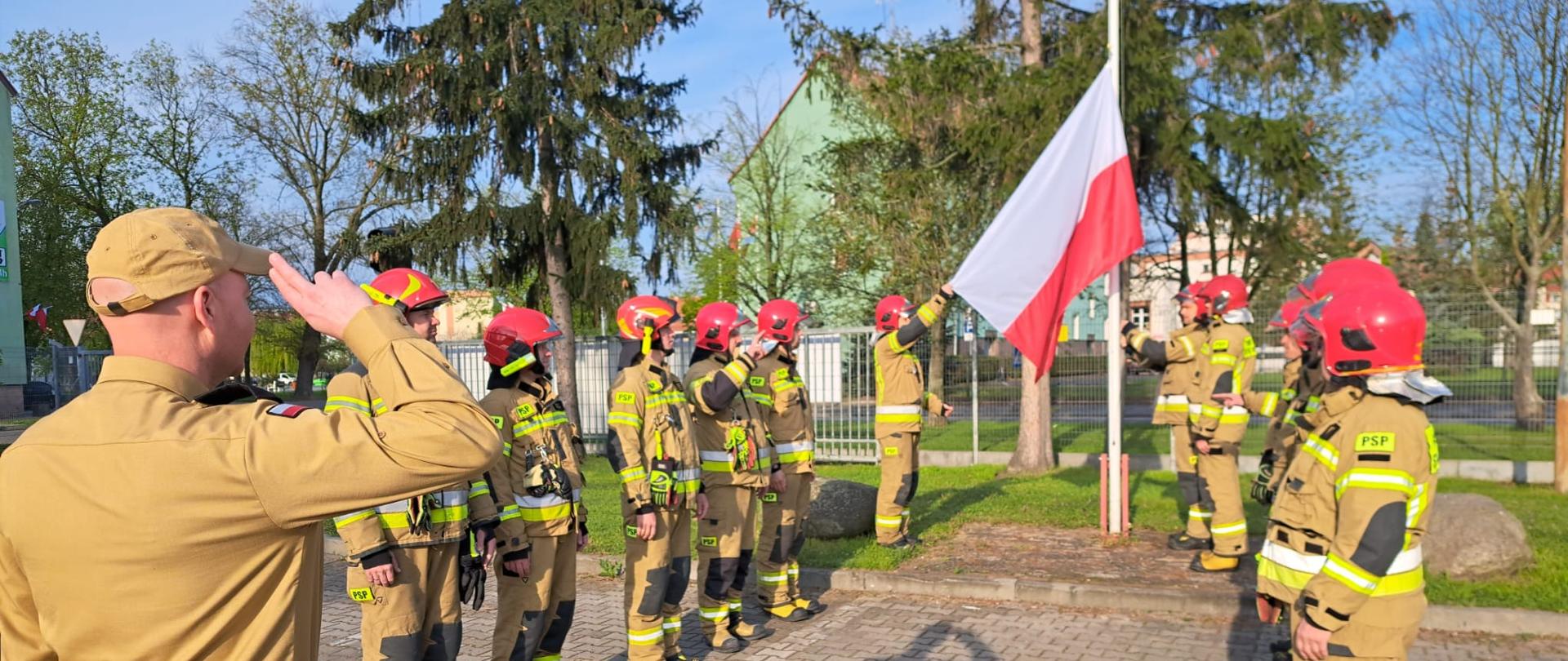 Umundurowani strażacy stojący w dwóch szeregach twarzami do siebie. W tle maszt na który wciągana jest flaga Rzeczypospolitej Polskiej.