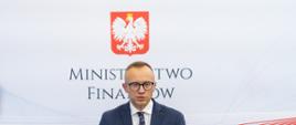 Wiceminister Artur Soboń podczas konferencji prasowej, w tle logo MF