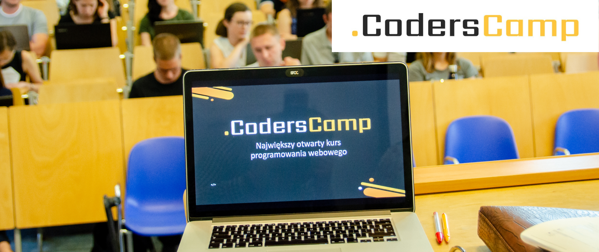 Grafika przedstawia laptopa, w którego tle widzimy salę wykładową. Na ekranie laptopa widoczny jest napis ".CodersCamp. Największy otwarty kurs programowania webowego." W prawym, górnym rogu widać napis brązowo-żółty ".CodersCamp."