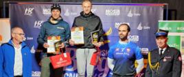 Zwycięzcy biegu stoją na podium obok organizatorzy biegu. Zwycięzcy prezentują nagrody. W tle ścianka okolicznościowa organizatora biegu. 