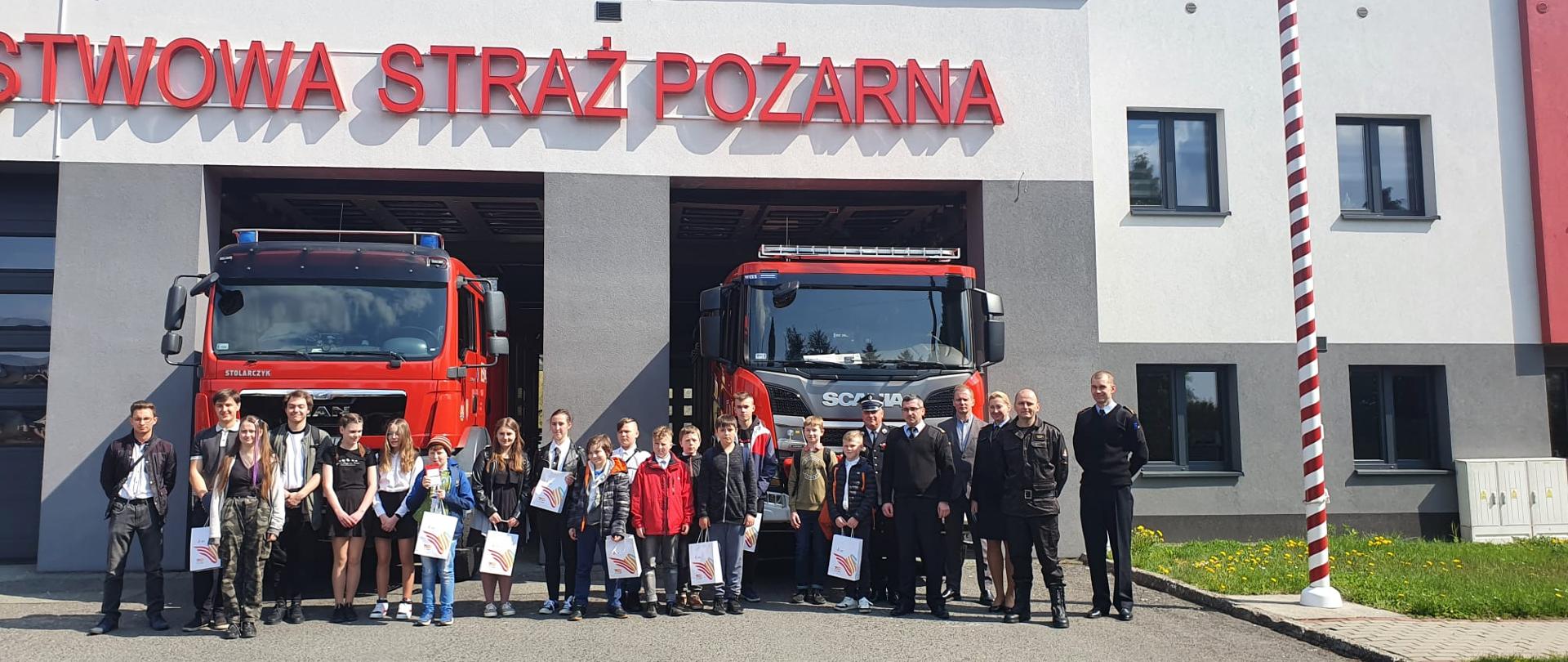 Uczestnicy turnieju stoją na tle dwóch samochodów pożarniczych i budynku komendy.
w rękach trzymają papierowe torby. w prawej części zdjęcia pracownicy Komendy.