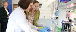 Minister funduszy i polityki regionalnej Katarzyna Pełczyńska-Nałęcz stoi przy stole laboratoryjnym na którym są fiolki i naczynia do badań, obok niej po prawej stronie stoi laborantka, a za plecami minister jest dyr. Międzynarodowego Instytutu Biologii Molekularnej i Komórkowej prof. Marta Miączyńska 
