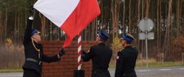 
Trzech strażaków w umundurowaniu galowym wciąga flagę państwową na maszt.
