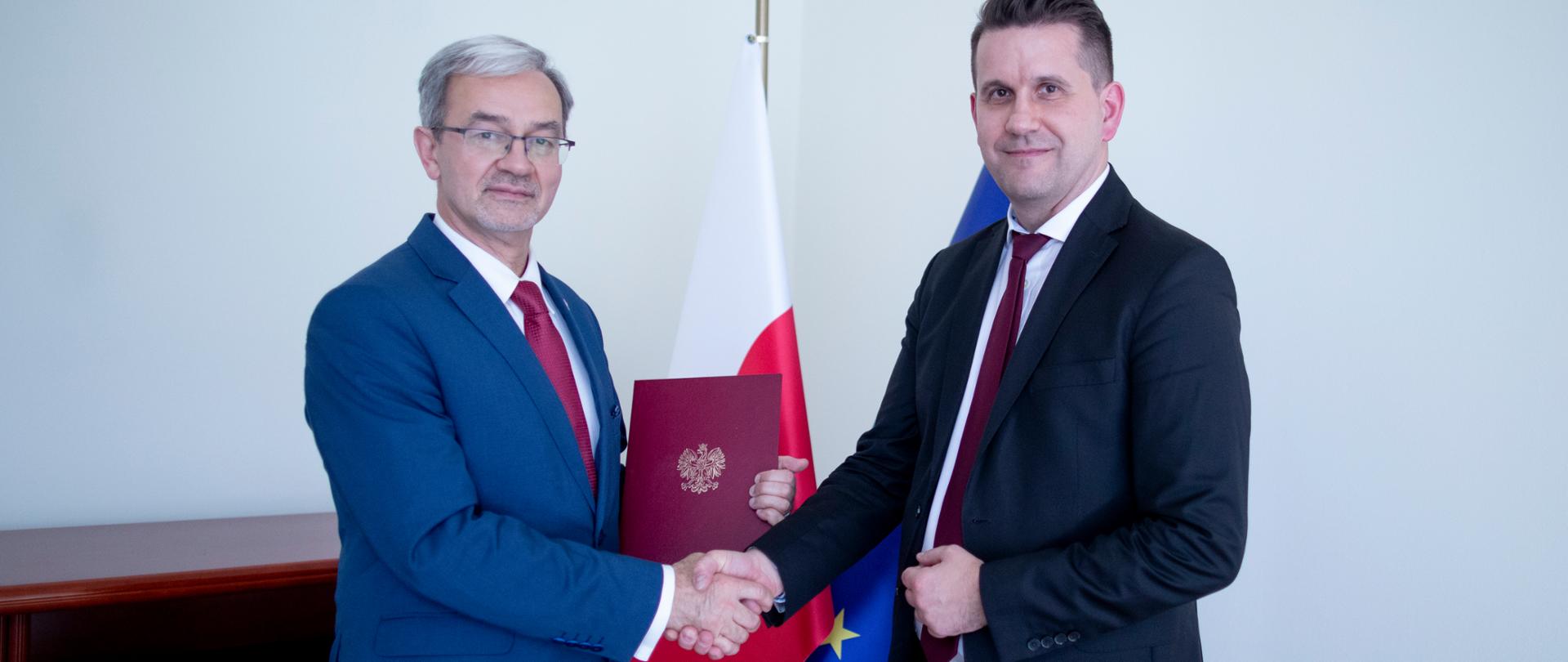 minister Jerzy Kwieciński i prezes Dominik Rozkrut ściskają prawe dłonie, między nimi widać teczkę z porozumieniem i flagi Polski i Unii Europejskiej