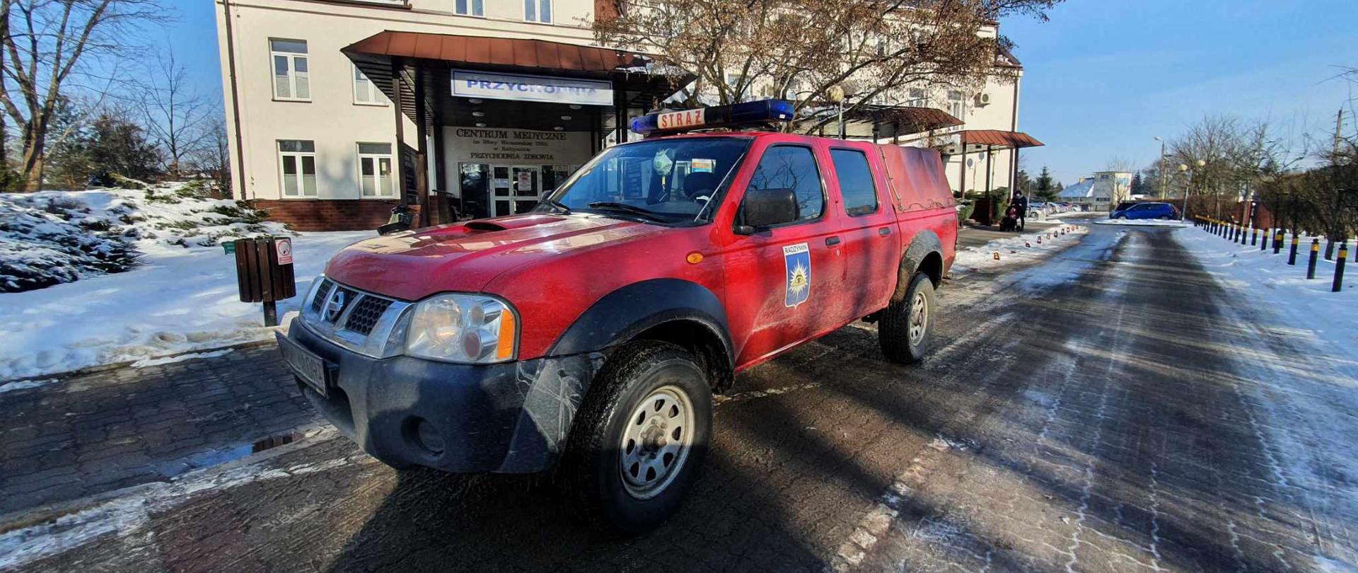 Na pierwszym planie na zdjęciu widać samochód OSP Radzymin typu pic-up. Za samochodem znajduję się budynek i wejście do przychodni, w której odbywają się szczepienia. Dookoła widać powoli topniejący śnieg.