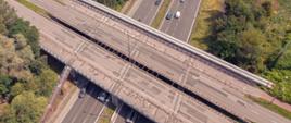 Widok z lotu ptaka na dwa wiadukty drogowe, przecinające się poniżej z inną drogą, na której widoczne są samochodów. Cień wiaduktu rzuca się na drogę poniżej, co wskazuje, że zdjęcie zostało zrobione w słoneczny dzień. Otoczenie dróg jest zielone.