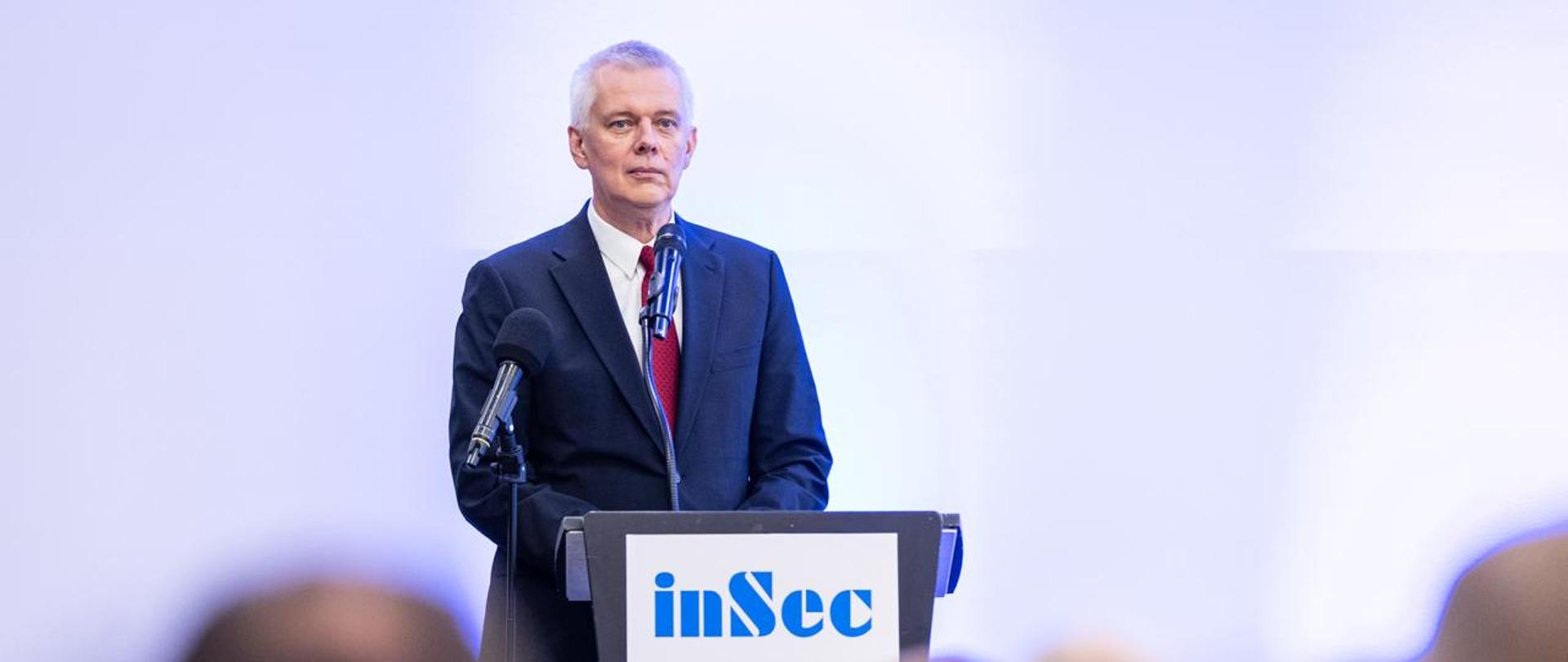 Minister Tomasz Siemoniak przemawia podczas konferencji InSec 2026. Minister stoi za szarą mównicą z napisem InSec (niebieskie litery na białym tle). Za mównicą dwa czarne mikrofony. Za ministrem białe tło. Na dole zdjęcia widać siedzących tyłem innych uczestników konferencji.