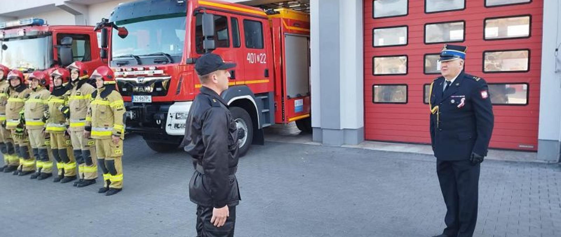 Zdjęcie wykonane przed budynkiem komendy. Centralny punkt stanowi komendant w galowym mundurze i stojący przed nim dowódca jednostki w mundurze sztabowym. W tle widać bramy garażowe oraz strażaków w mundurach piaskowych i czerwonych hełmach. Strażacy stoją w rzędzie i słuchają meldunku dowódcy dla komendanta.