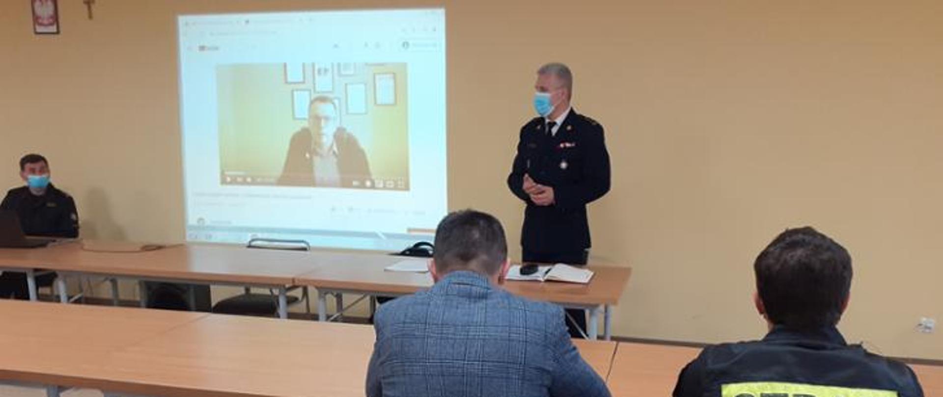 Komendant powiatowy zwraca się do słuchaczy. W tle prezentacja.