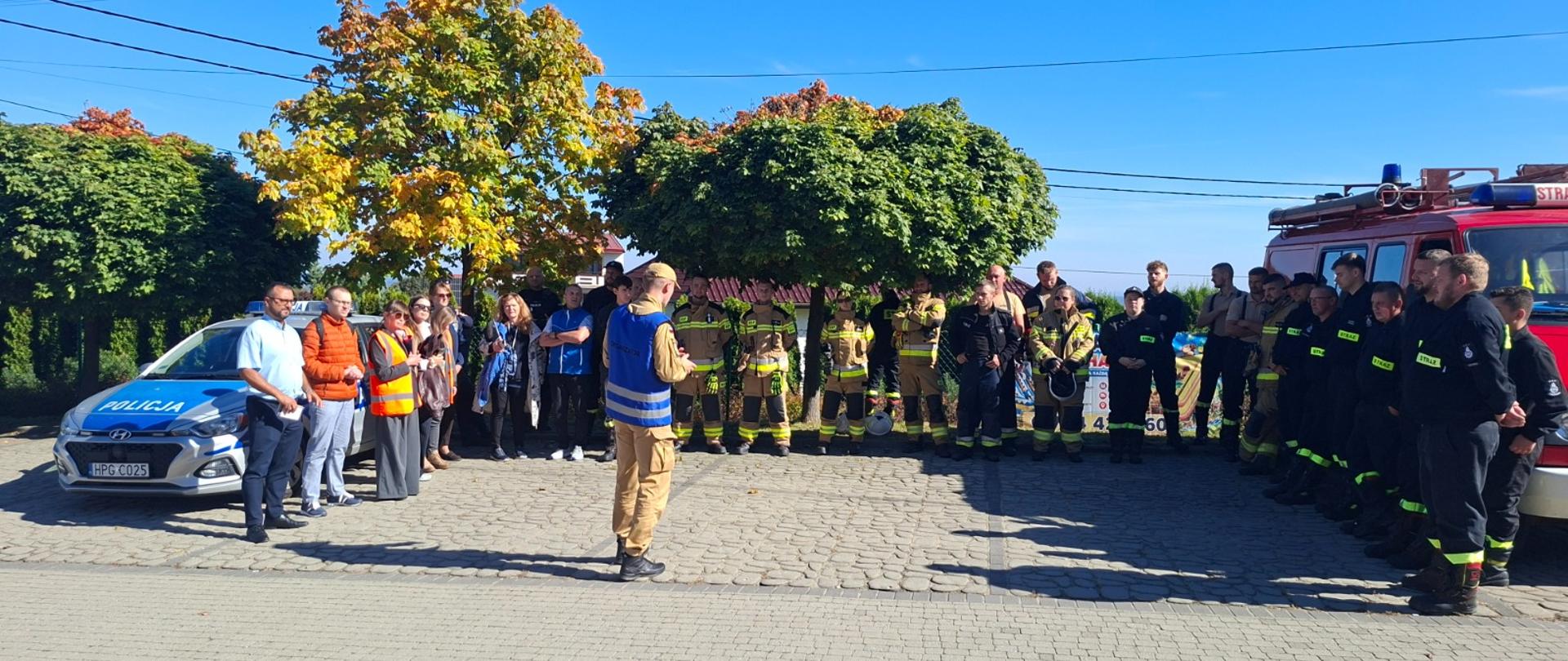 Na placu stoją w podkowie osoby biorące udział w ćwiczeniach bo jednej stronie stoi radiowóz policyjny z drugiej strony wóz strażacki. W oddali widać drzewa.