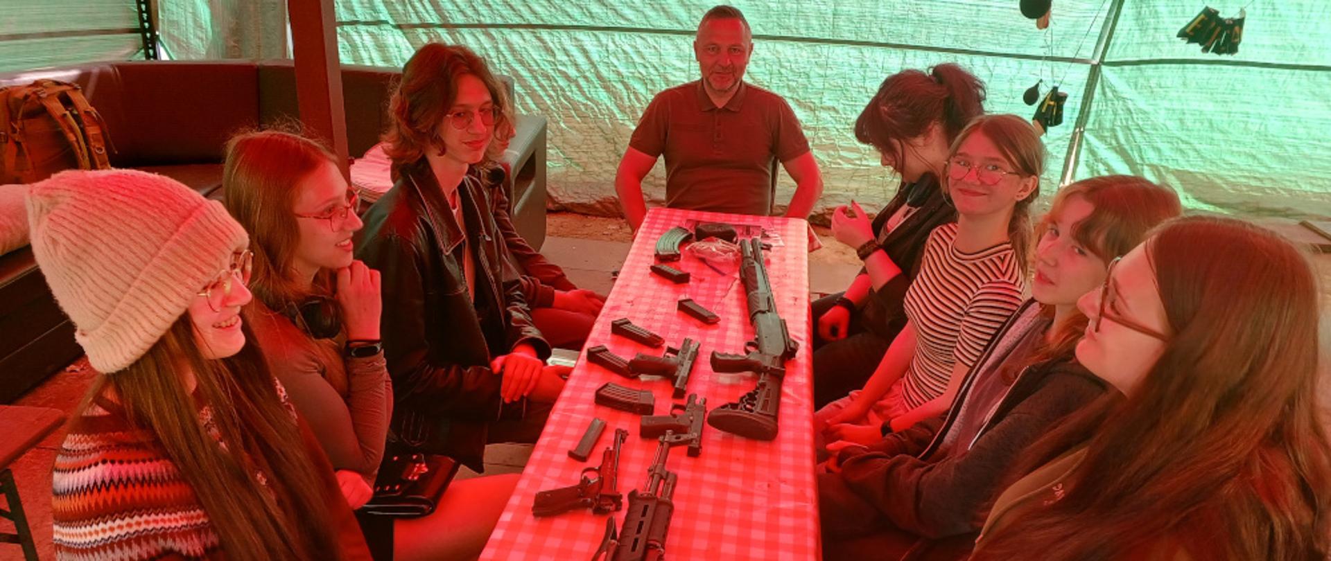 Uczniowie ZPSM siedzą przy stole, na którym porozkładana jest broń palna, na końcu siedzi instruktor