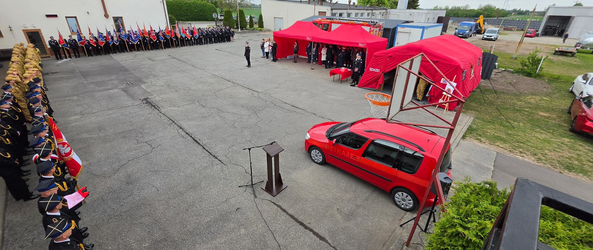 Widok z góry na plac uroczystości z pocztami sztandarowymi, strażakami ustawionymi w szyku oraz czerwonymi namiotami przygotowanymi na obchody.