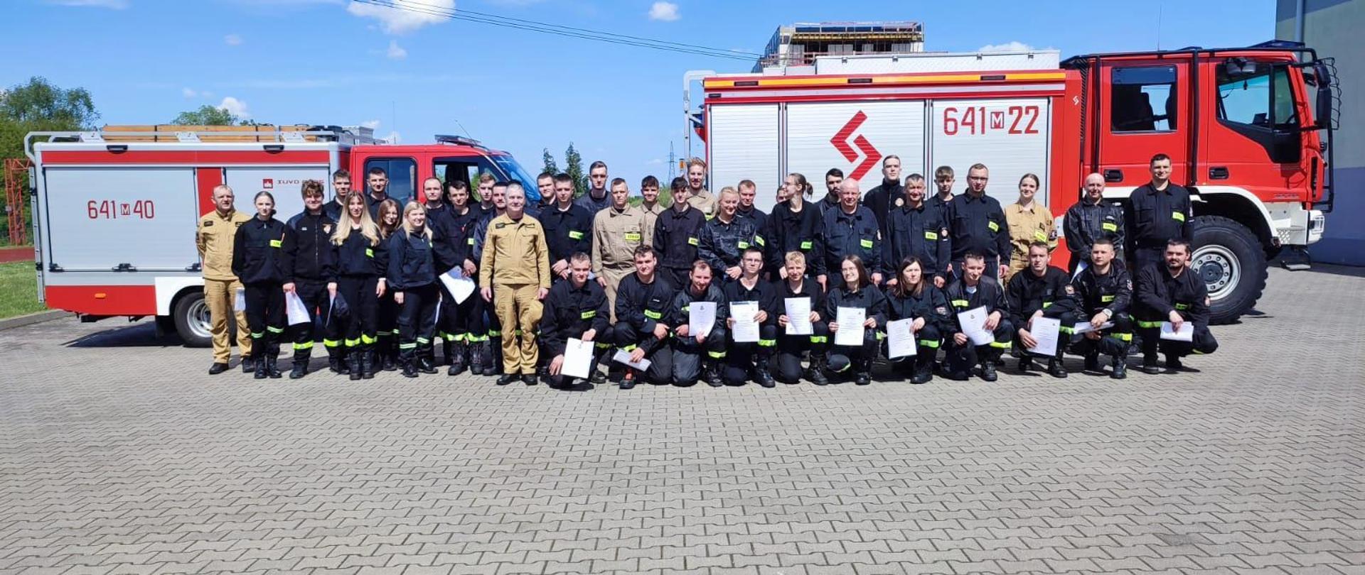 Egzamin końcowy szkolenia podstawowego strażaków ratowników Ochotniczych Straży Pożarnych - pamiątkowe zdjęcie na tle pojazdów pożarniczych.