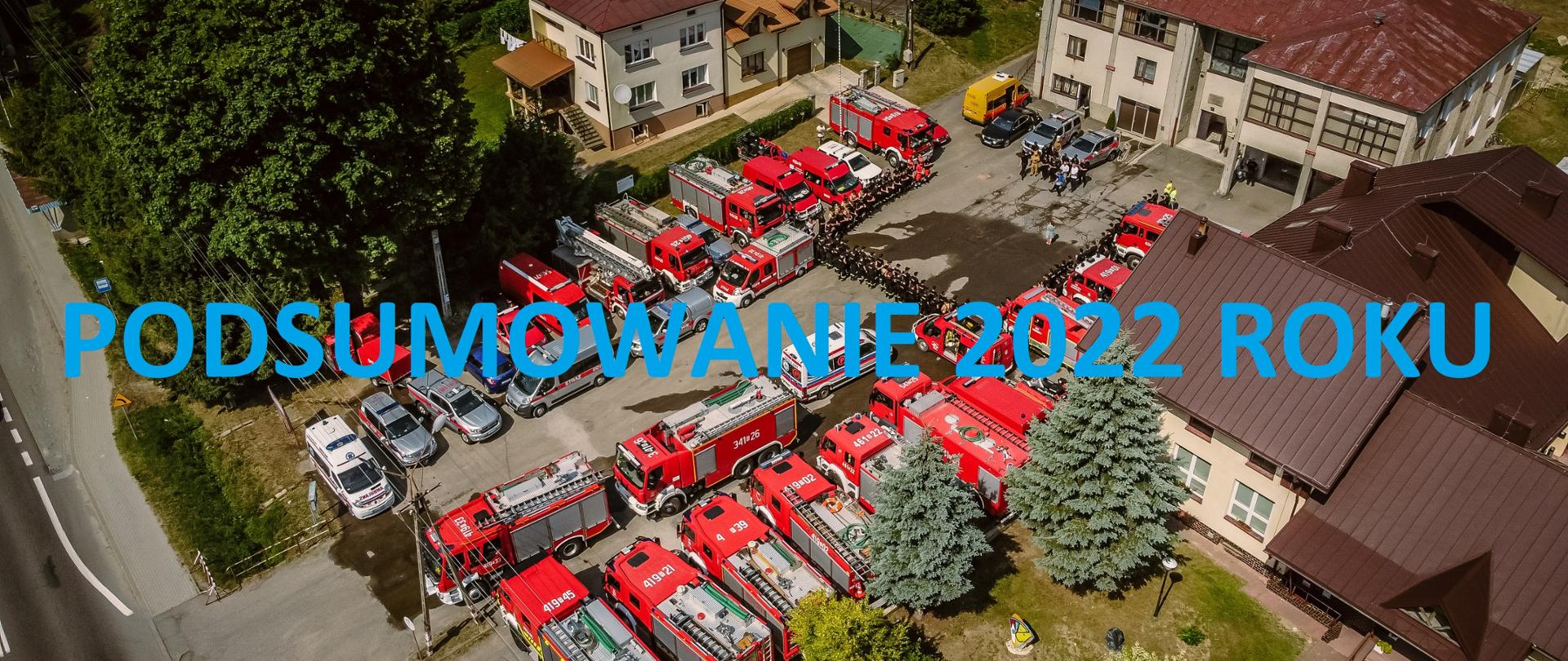Kolorowa fotografia wykonana w słoneczny dzień na zewnątrz. Przedstawia ona z lotu ptaka miejsce zbiórki strażaków i czerwonych ratowniczych na placu przed remizą w czasie ćwiczeń strażackich. Na zdjęciu niebieski napis posumowanie 2022 roku.