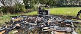 Spalony pojazd dostawczy oraz rozrzucone na polu pozostałości po pożarze. W tle widoczne drzewa i teren rolniczy.